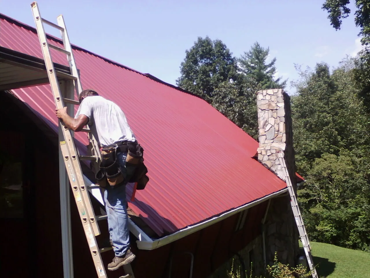Roofer on a ladder beside a red metal roof and stone chimney — Soffit & Fascia Repair by Roofing Brothers in Altair